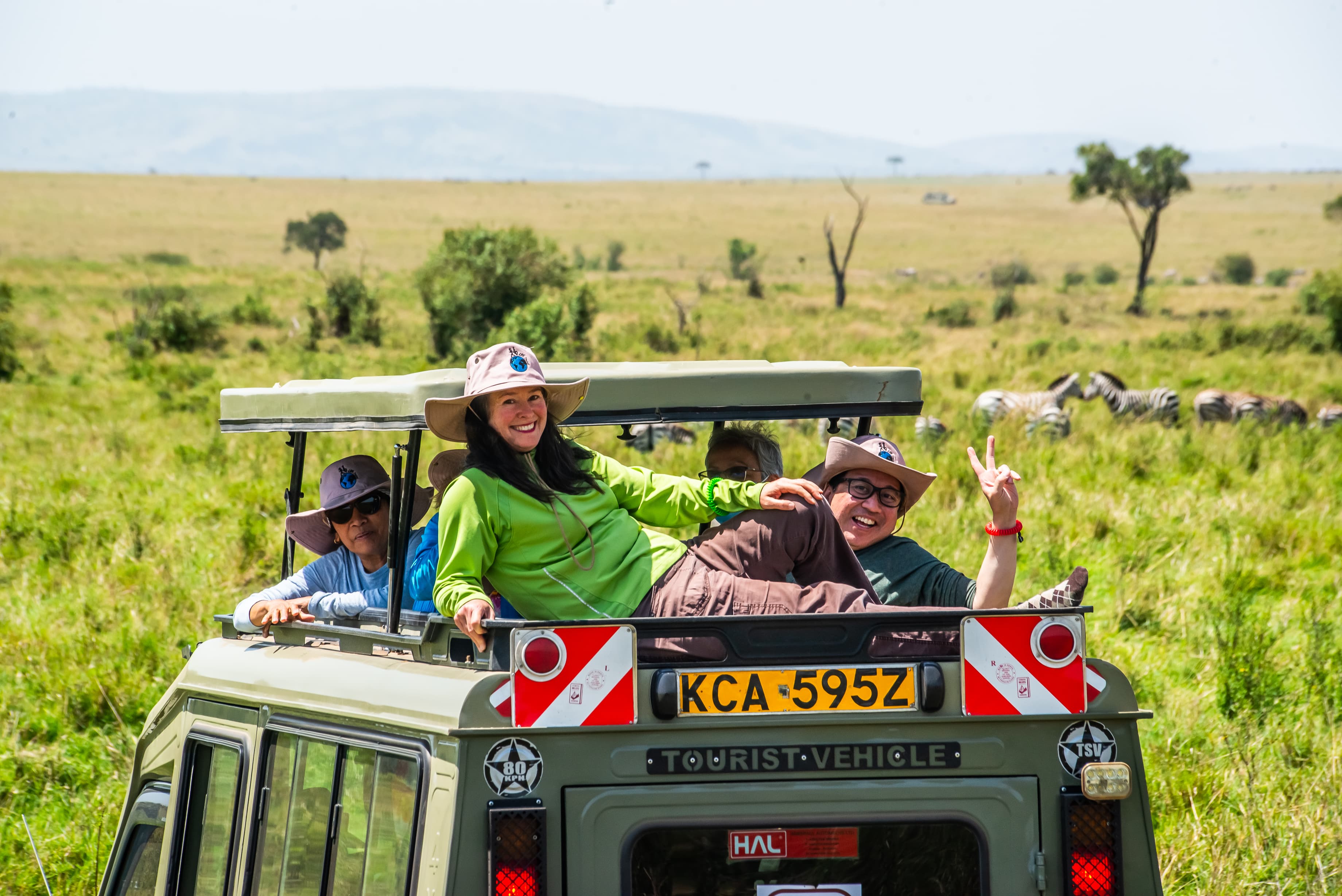 Guests standing through the roof hatch of a Kenya safari vehicle with zebras grazing on the Masai Mara plains in the background