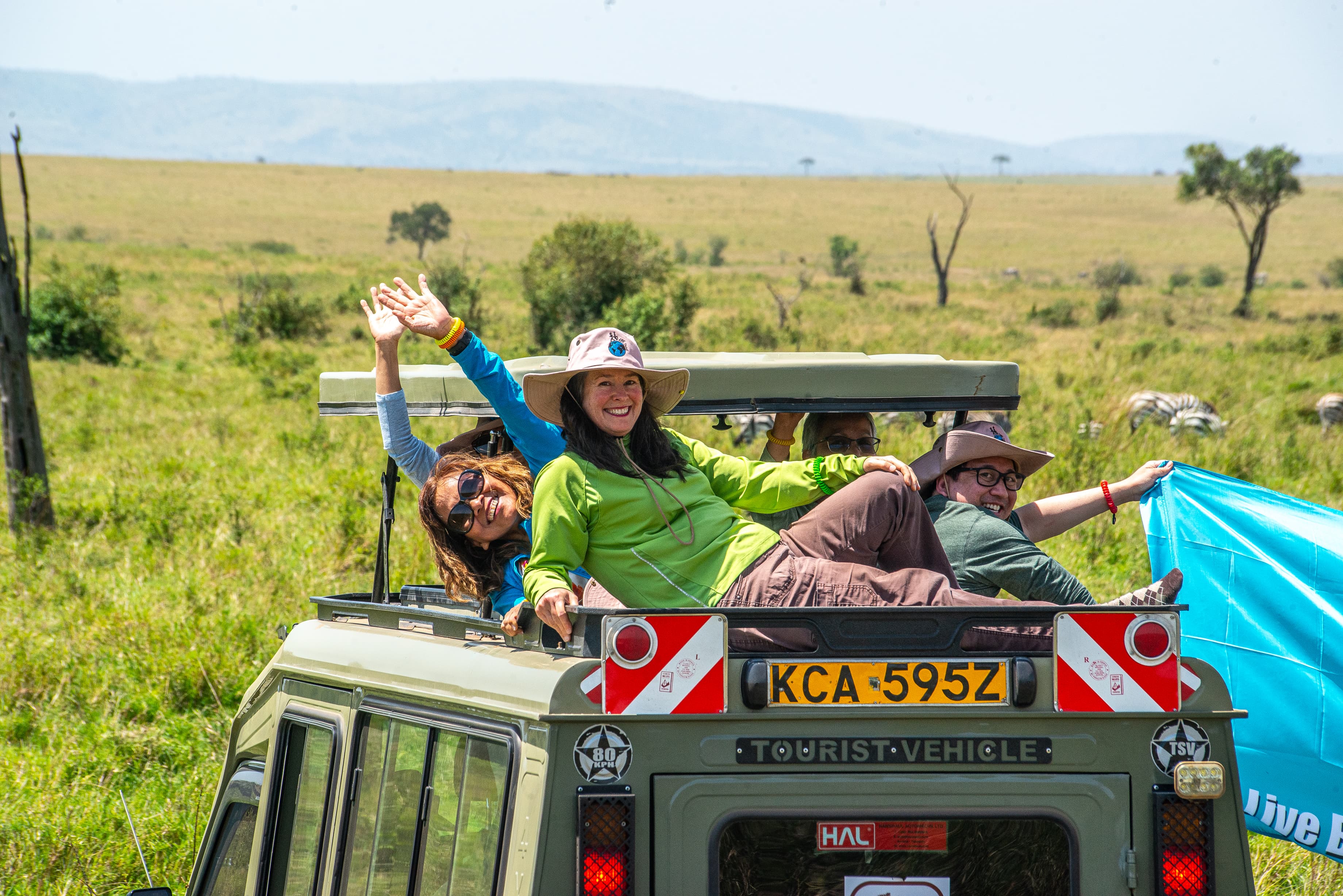 Happy guests on a pop-top game drive through the Mara grasslands &mdash; zebra graze indifferently in the background. This is what it actually looks and feels like out there.
