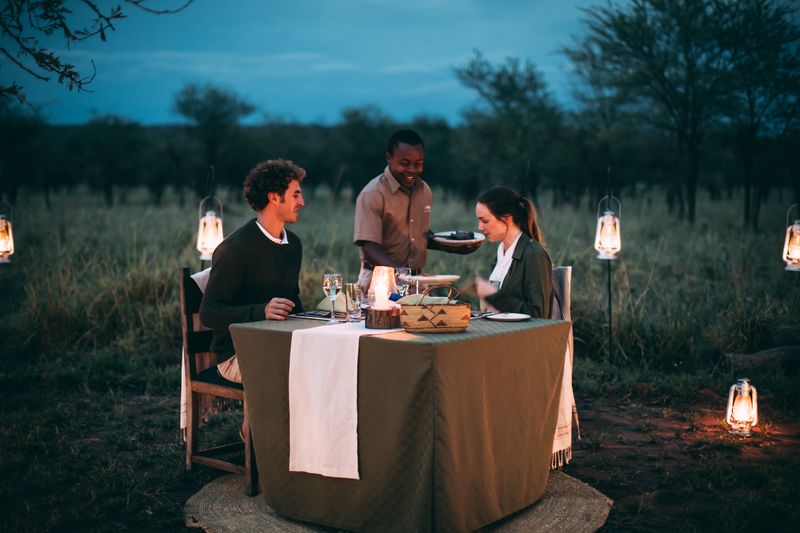 Couple enjoying a romantic bush dinner under lantern light, served by staff in a peaceful safari setting at dusk.